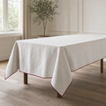 A rectangular chalk-white linen tablecloth with a cherry-red bound edge and hand-run stitch line draped over a dining table