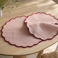 Two round blush-pink linen placemats with a scalloped cherry-red bound edge and hand-run stitch line on a pale oak dining table