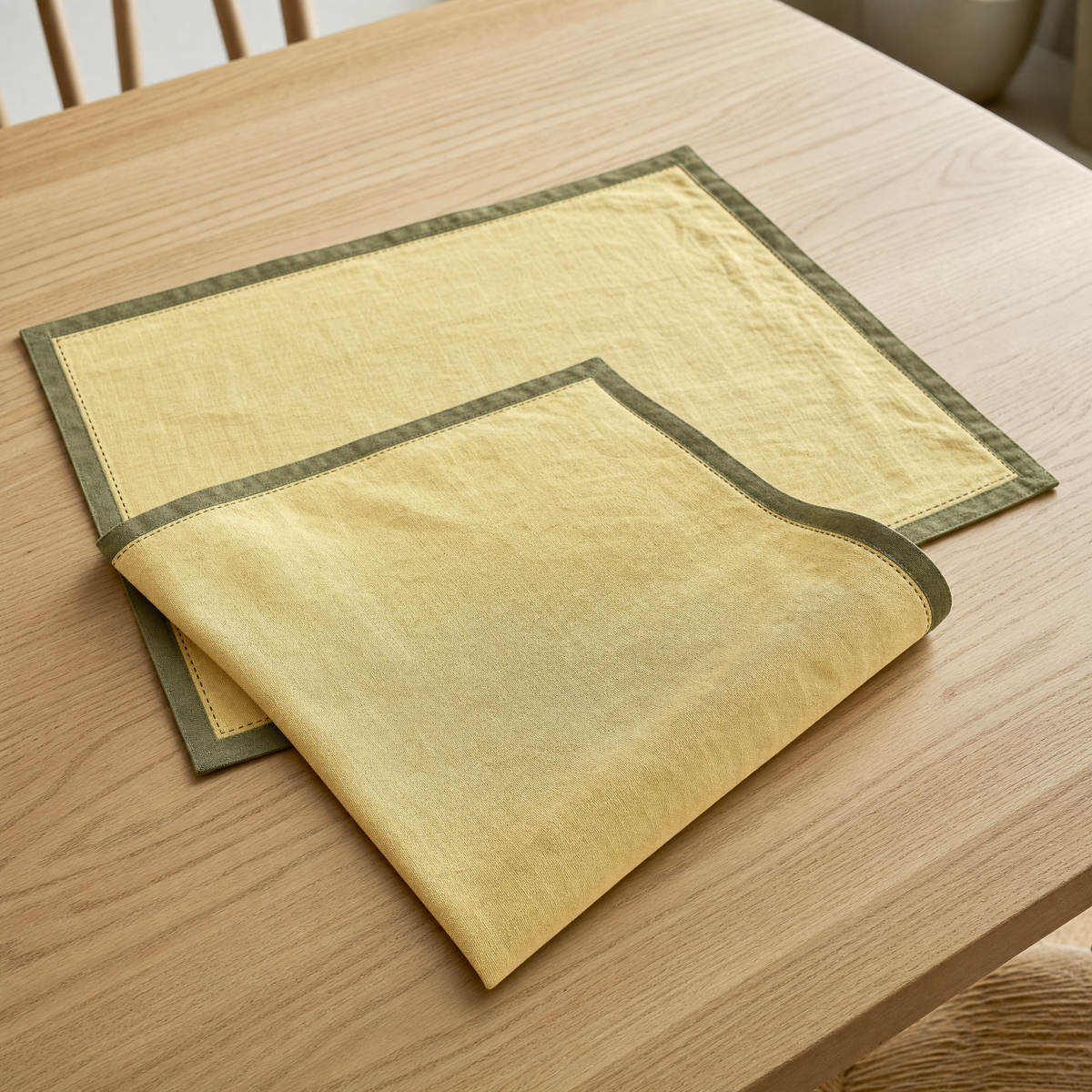 Two rectangular butter-yellow linen placemats with an olive-green bound edge and hand-run stitch line on a pale oak dining table